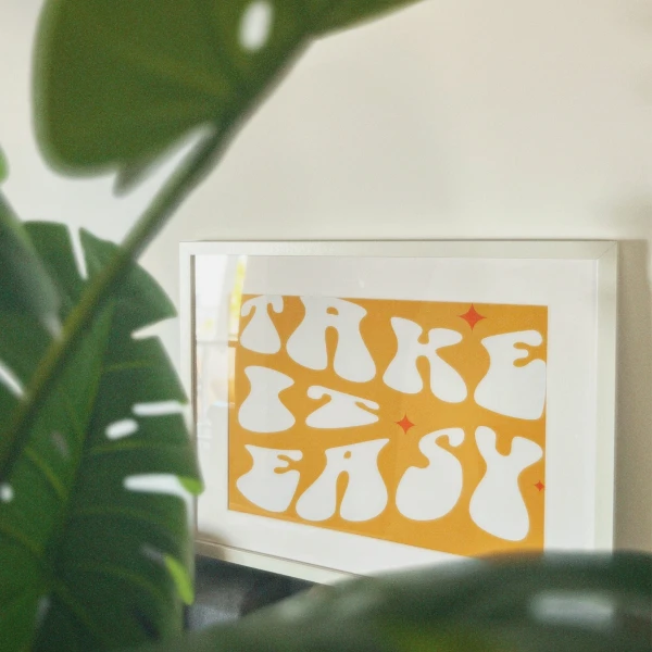 Framed artwork with white text 'MAKE IT EASY' on an orange background, partially obscured by green plant leaves.