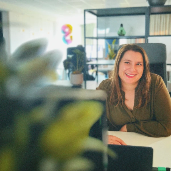 Smiling woman seated at a desk in a modern office with plants and shelves in the background.