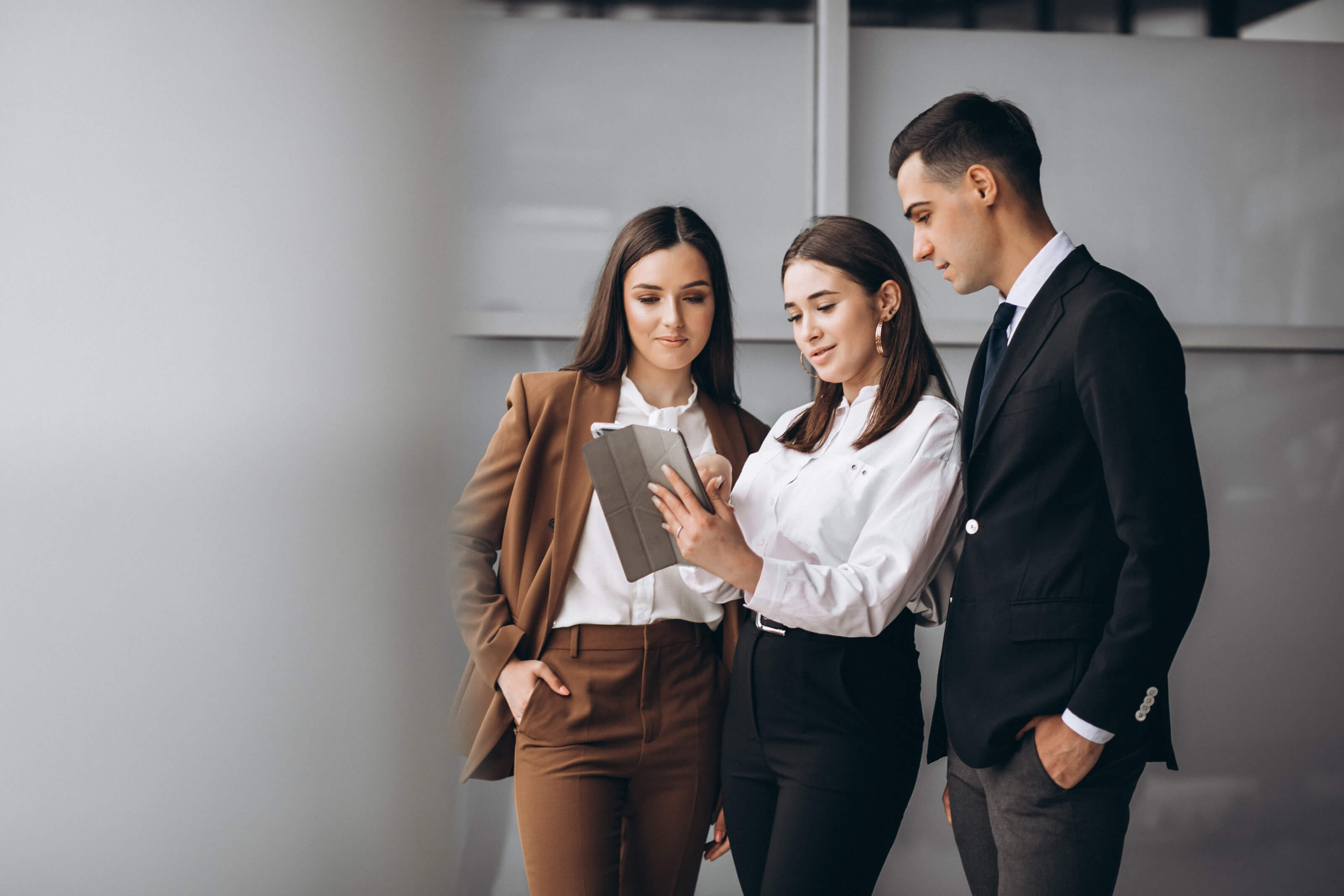 Three professionals standing together in a modern office environment, reviewing information on a digital tablet held by the woman in the center.