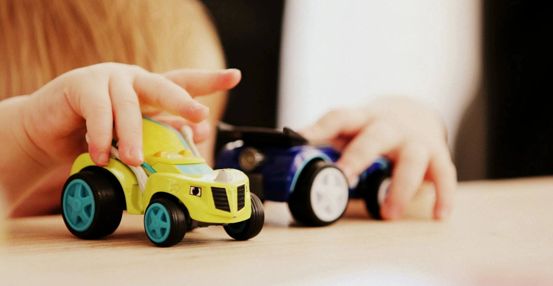 Close-up of a child's hands pushing a yellow toy car next to a blue toy car on a wooden surface.