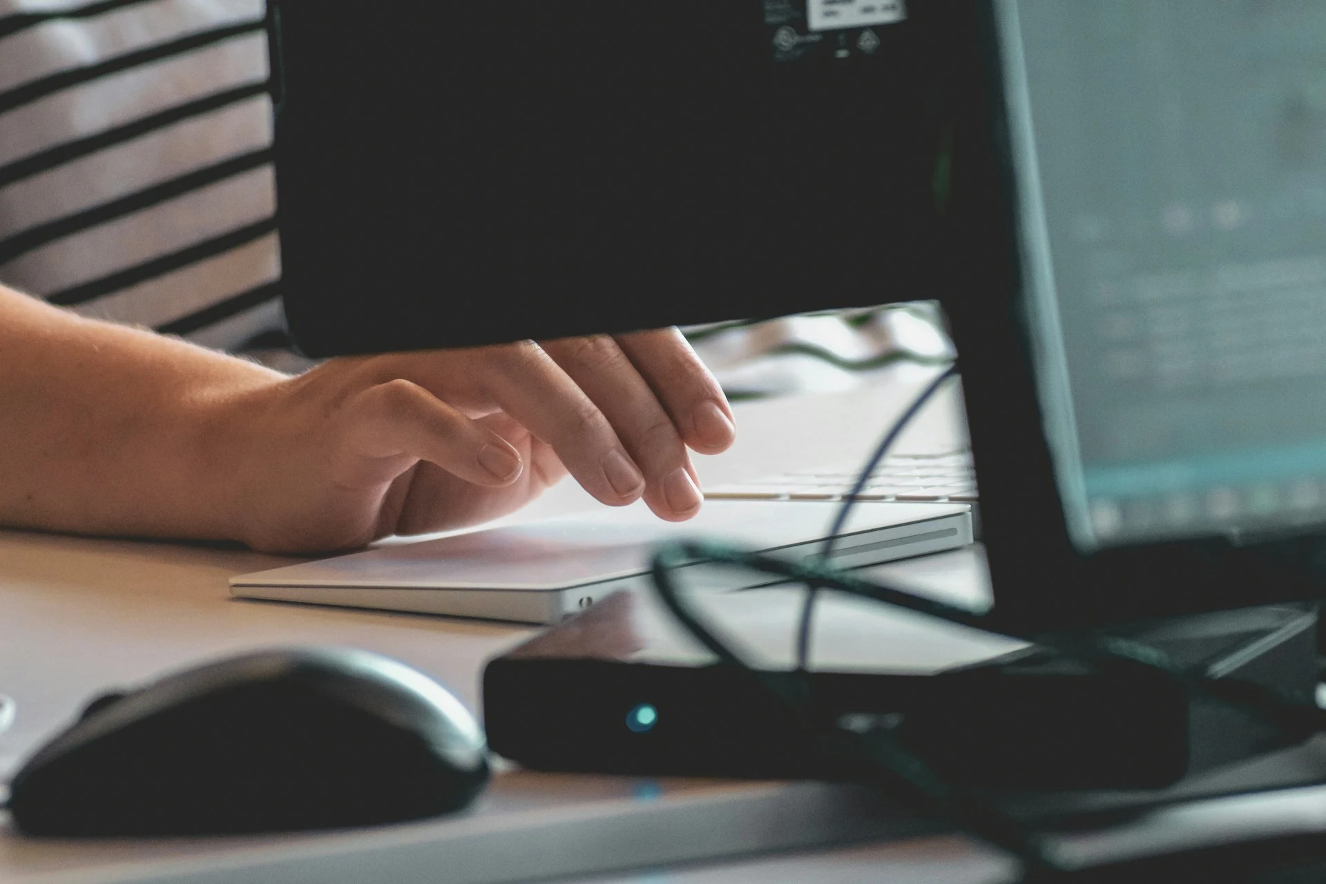 Close-up side view of a professional's hand navigating a laptop trackpad in a workspace, with a computer monitor frame in the foreground.