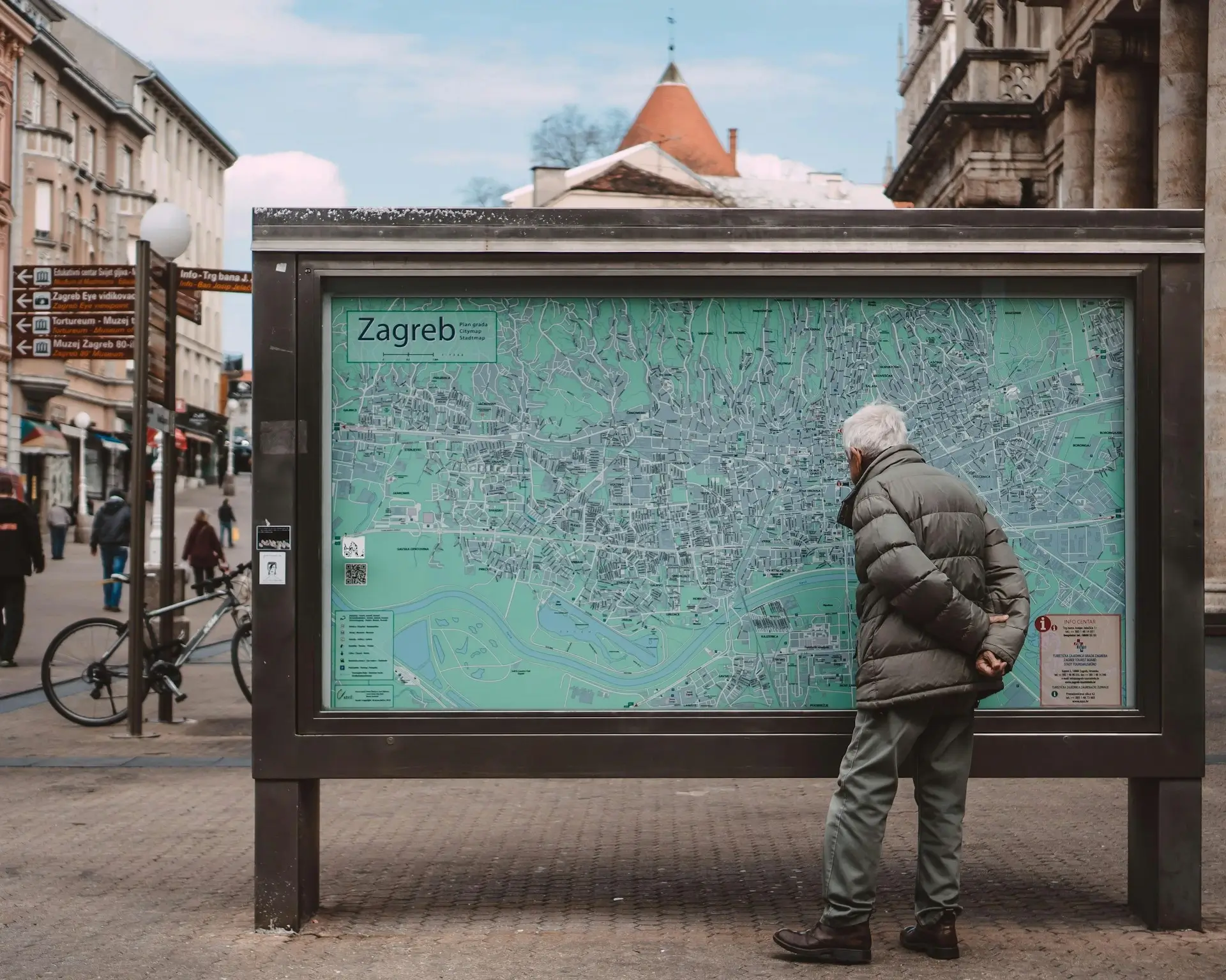 Rear view of an older gentleman standing on a city sidewalk, examining a large, green public map of Zagreb displayed in a freestanding glass case.