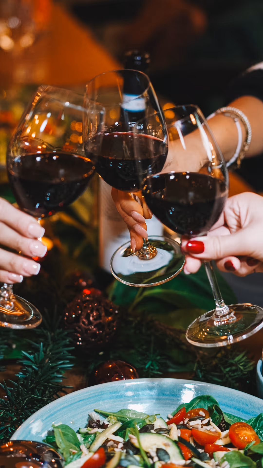 Drei Hände stossen mit Gläsern Rotwein über einem Tisch an, auf dem ein frischer Salat und Weihnachtsschmuck stehen.