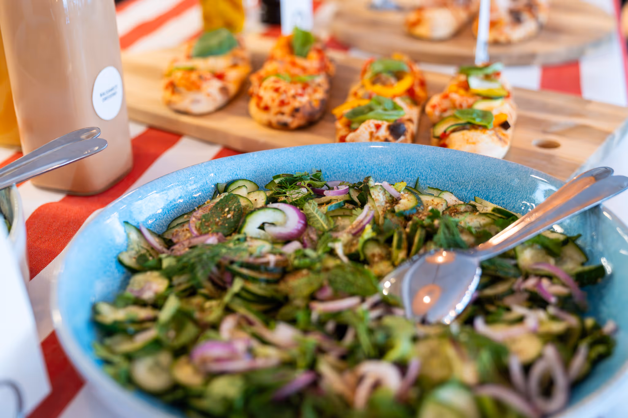 Eine blaue Schüssel, gefüllt mit frischem Gurkensalat mit roten Zwiebeln und Kräutern, im Hintergrund Pizza Bites.