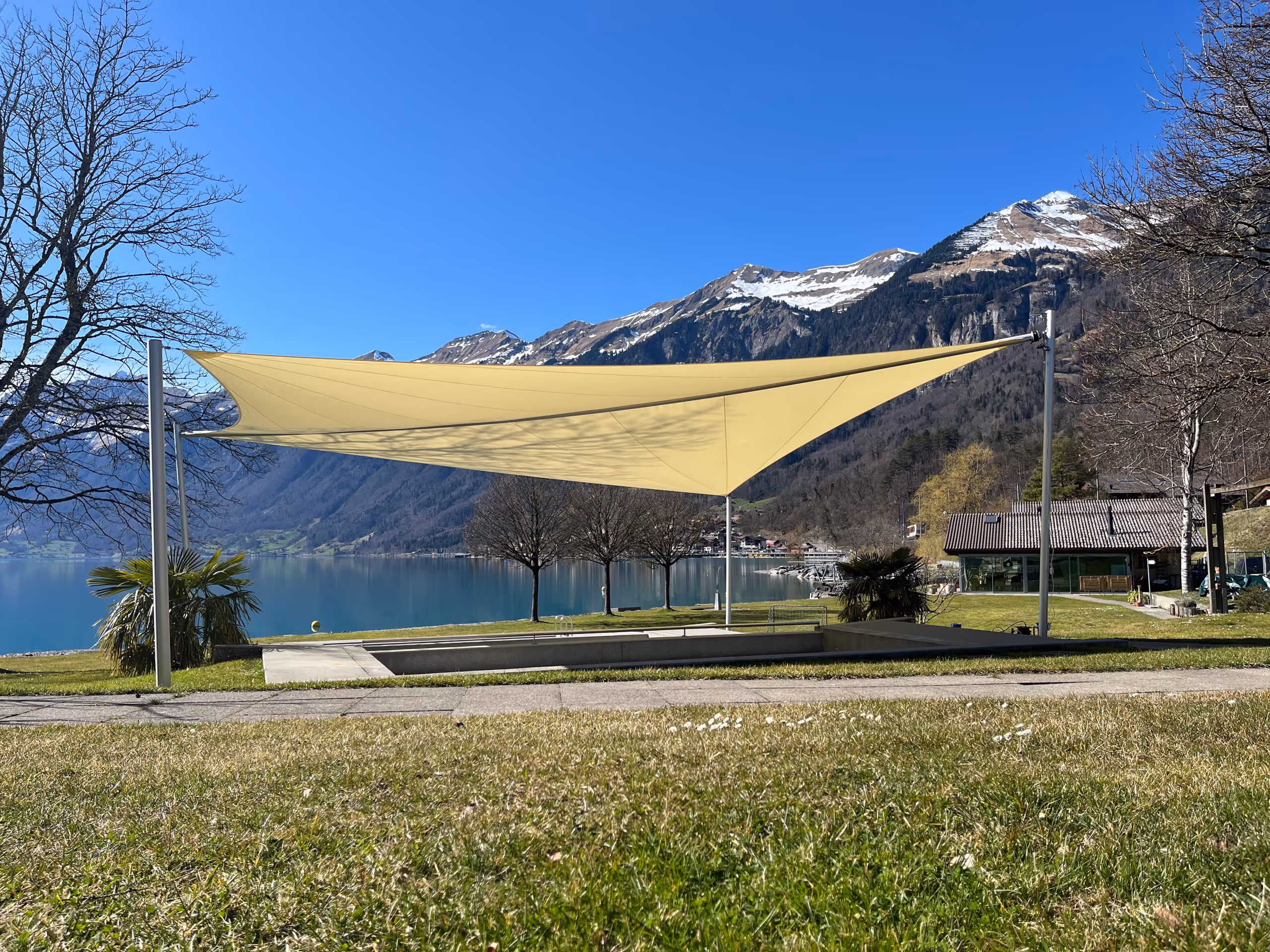 Gelbes Sonnensegel über einem leeren Pool in einer Wiese mit Blick auf See und schneebedeckte Berge im Hintergrund.