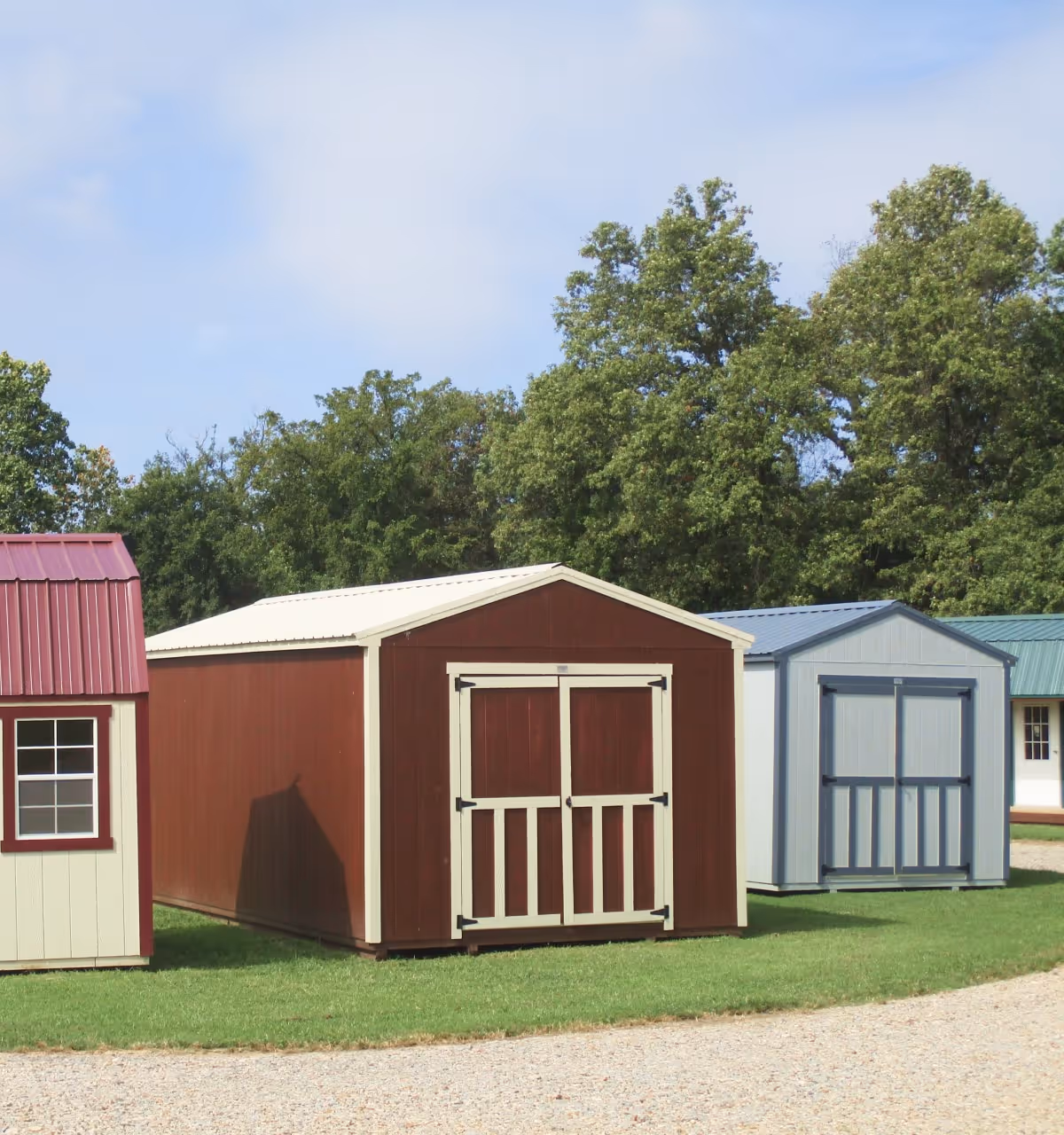 Wooden storage sheds with different roof colors placed on grass near a gravel path, with trees in the background.