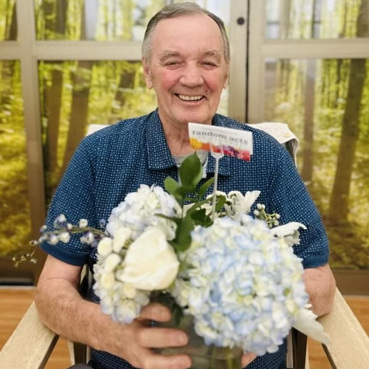 A man sitting smiles warmly while holding a vase of fresh flowers, including hydrangeas, with a small Random Acts of Flowers card placed in the arrangement.