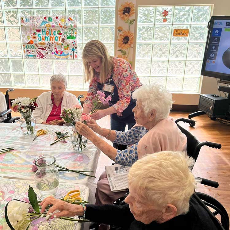 A volunteer in a pink shirt and purple apron leans over a table. At the table there are three older women who are building their own bouquets.