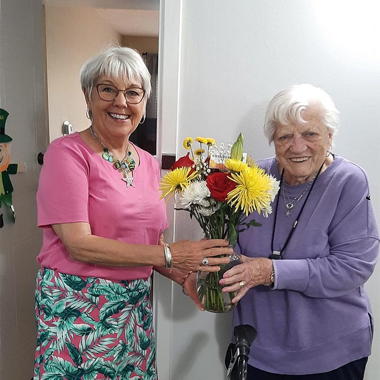 Two women stand side by side. The woman on the left wears a pink shirt and has short gray hair. The woman on the right wears a purple shirt and has short white hair. Together, they hold a vase filled with flowers positioned between them.