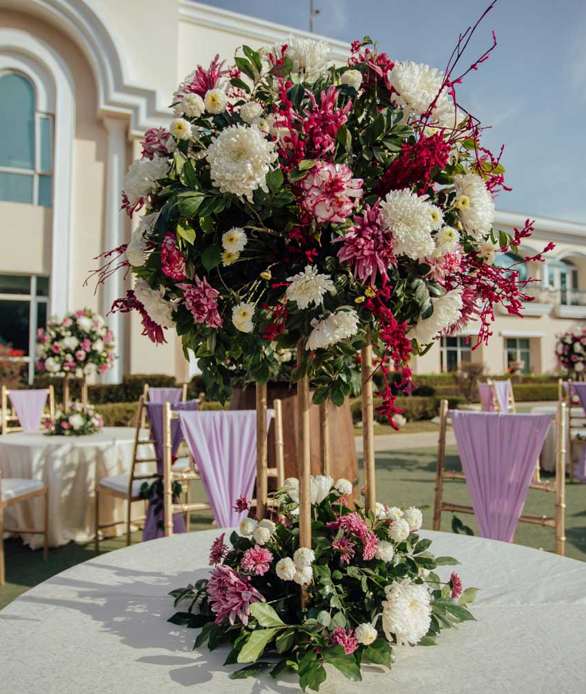 A large sphere bouquet of flowers stands above a white circular table. At the base of the stand holding the large flower sphere, there is another bouquet. The flowers have dark green leaves with light and dark pink and cream colored flowers.