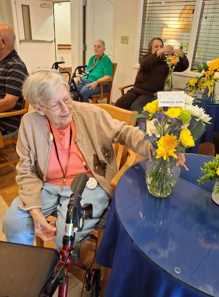 A circular table with a navy blue tablecloth sits to the right, accompanied by an older woman in a wheelchair wearing a coral and tan shirt to the left. The woman is reaching for a glass vase with a yellow bouquet.