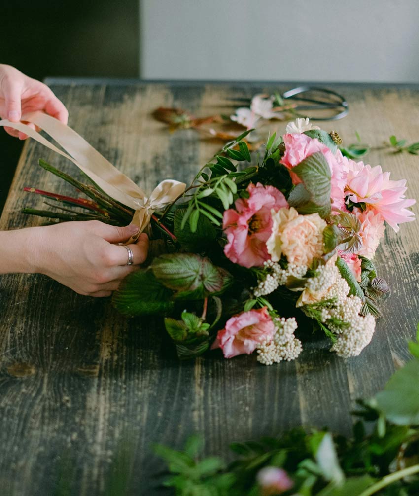 A bouquet of flowers, tied with a cream-colored ribbon, lies on a wooden table. The flowers are a mix of pale and more vibrant pink, with some smaller white ones mixed in.