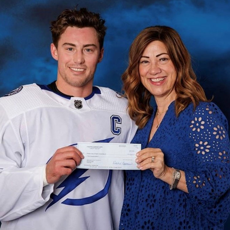 A man and woman stand in front of a marbled blue background. They are both holding onto a check that is placed between them. The man is in a white hockey jersey, and the woman is in the royal blue top. 