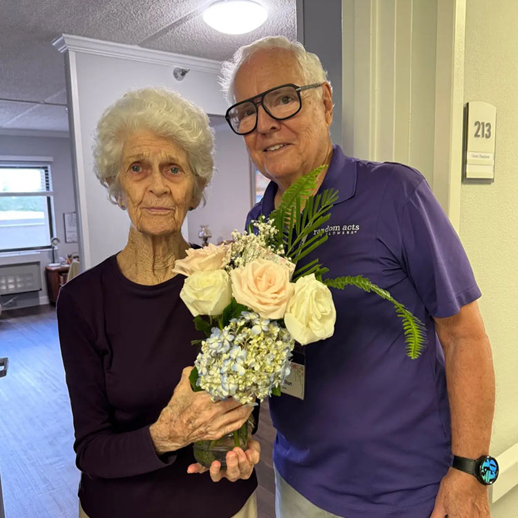 An older woman and a man stand together posing for the camera. They are both wearing purple shirts. The woman is holding a white bouquet of flowers.