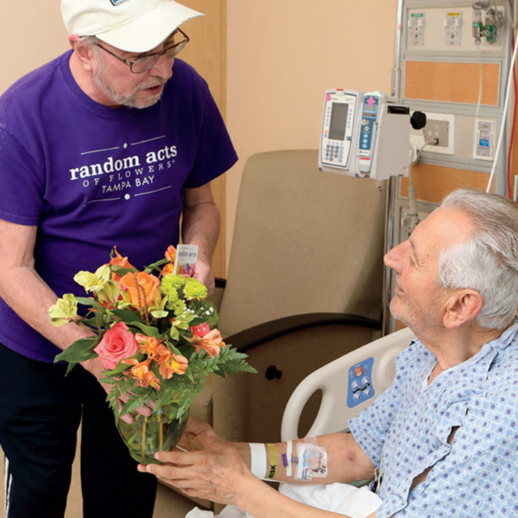 A Random Acts of Flowers volunteer in a purple shirt delivers a bright bouquet to a hospital patient, who smiles warmly while accepting the arrangement.