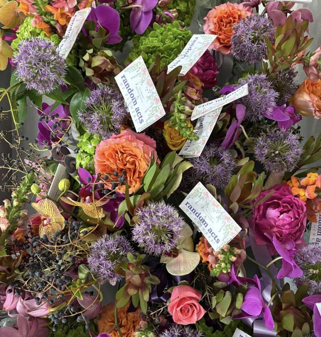 A close-up of several floral arrangements. Each arrangement features a small Random Acts of Flowers card. Colors of the flowers include coral, light and dark pink, purple, and some green.
