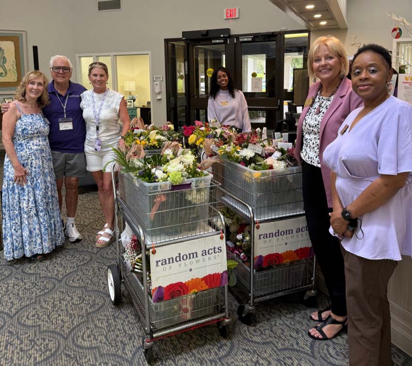 Two metal carts, each with a sign that reads “Random Acts of Flowers,” are filled with flowers. About four volunteers stand around the carts, smiling at the camera.