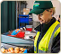 Man wearing a green cap, yellow safety vest, and black jacket standing near boxes in a warehouse or storage area.
