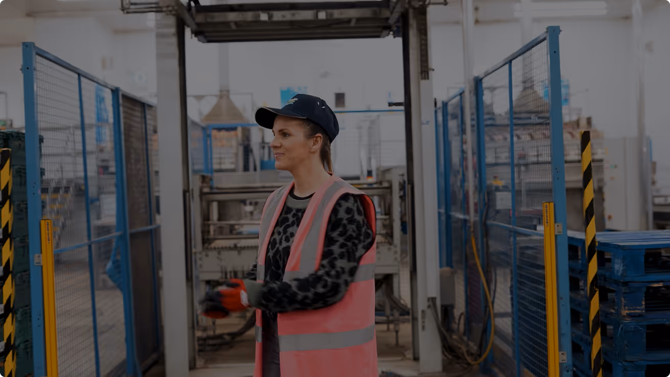 Woman wearing a pink safety vest and black cap standing in a factory or warehouse setting.