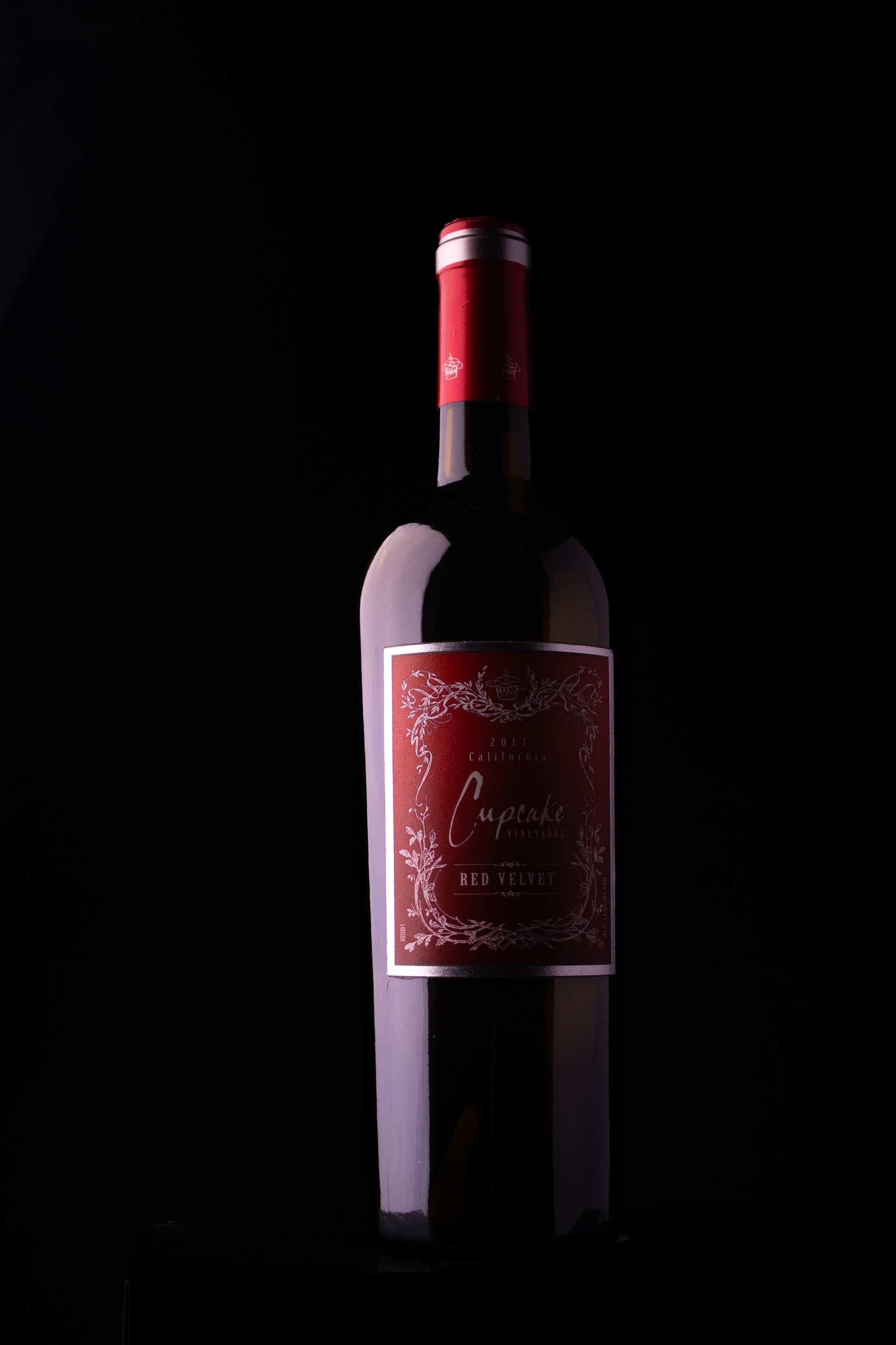A bottle of Cupcake Vineyards Red Velvet wine with a red label and red foil on the neck, set against a black background.