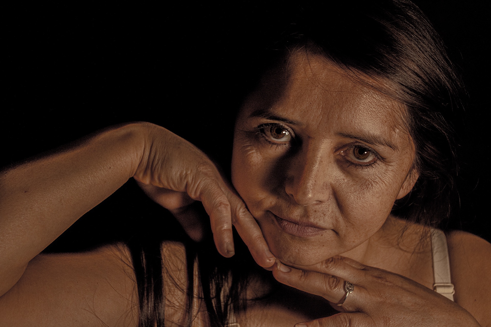 Close-up portrait of a woman with brown eyes resting her face on her hands against a dark background.