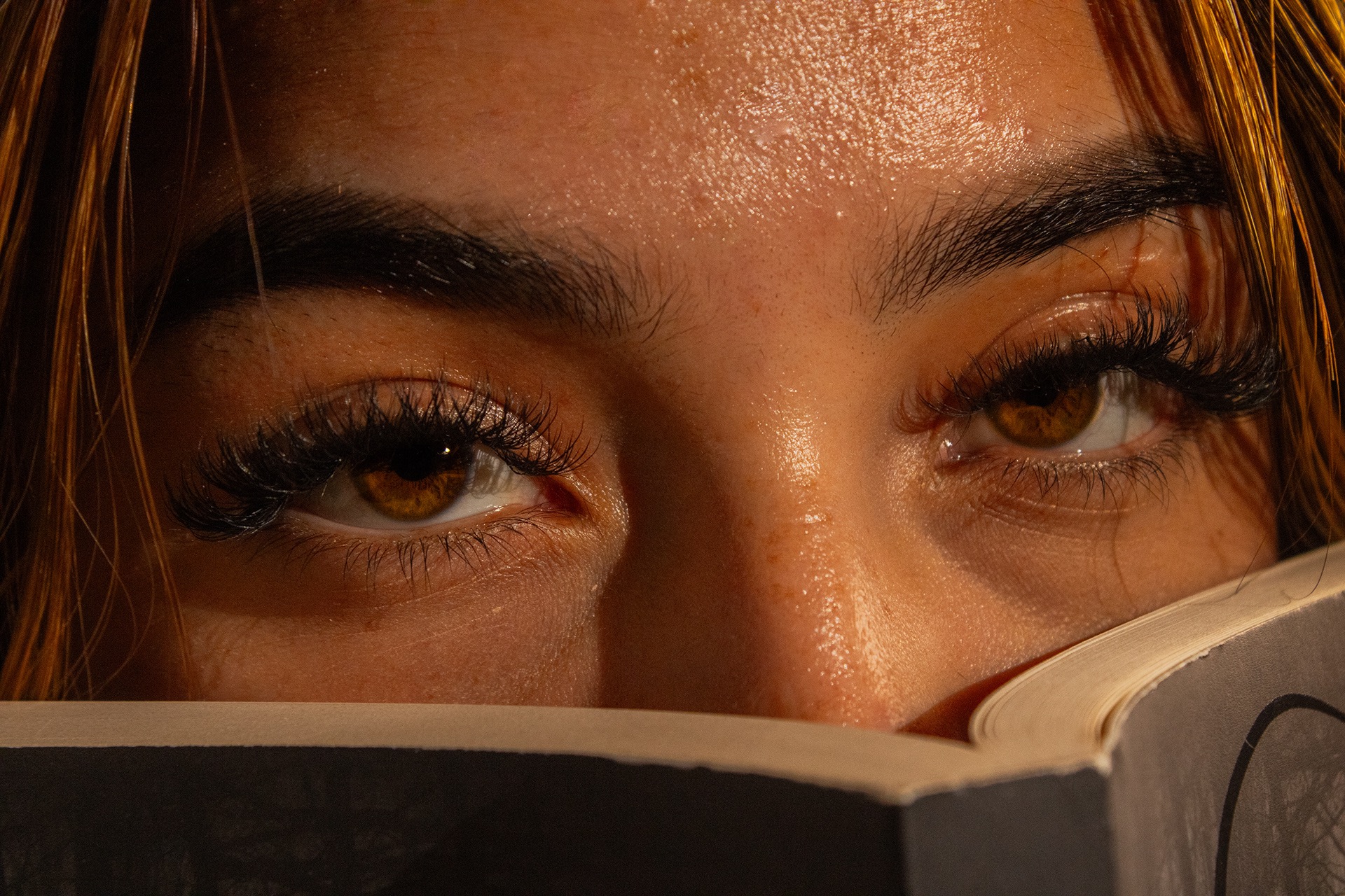 Close-up of a person's brown eyes and long eyelashes peeking over the top edge of a book.