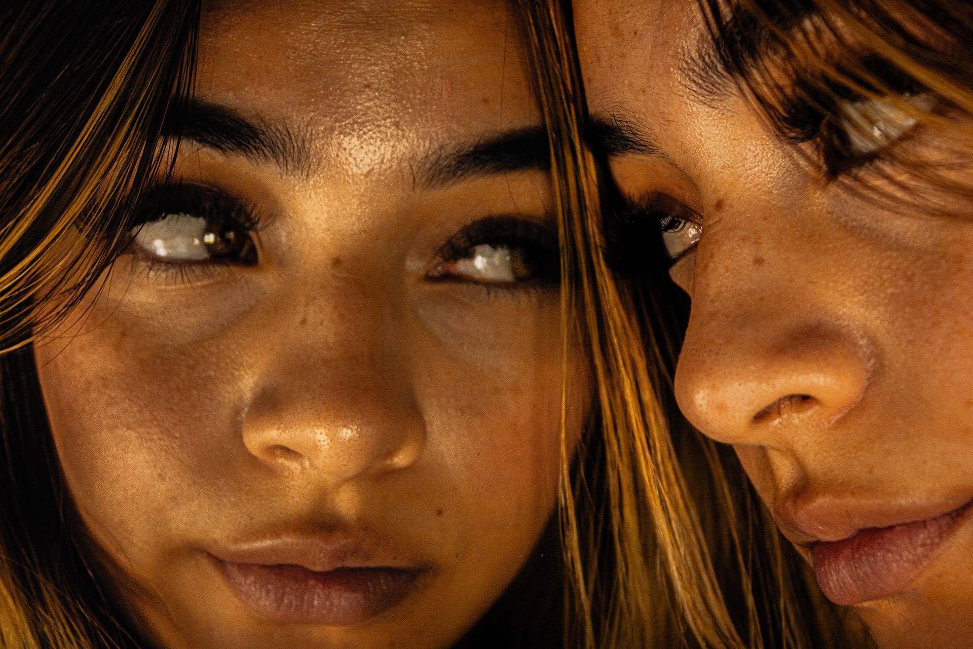 Close-up of two women's faces looking sideways towards each other, showing their eyes, noses, and lips with warm lighting.