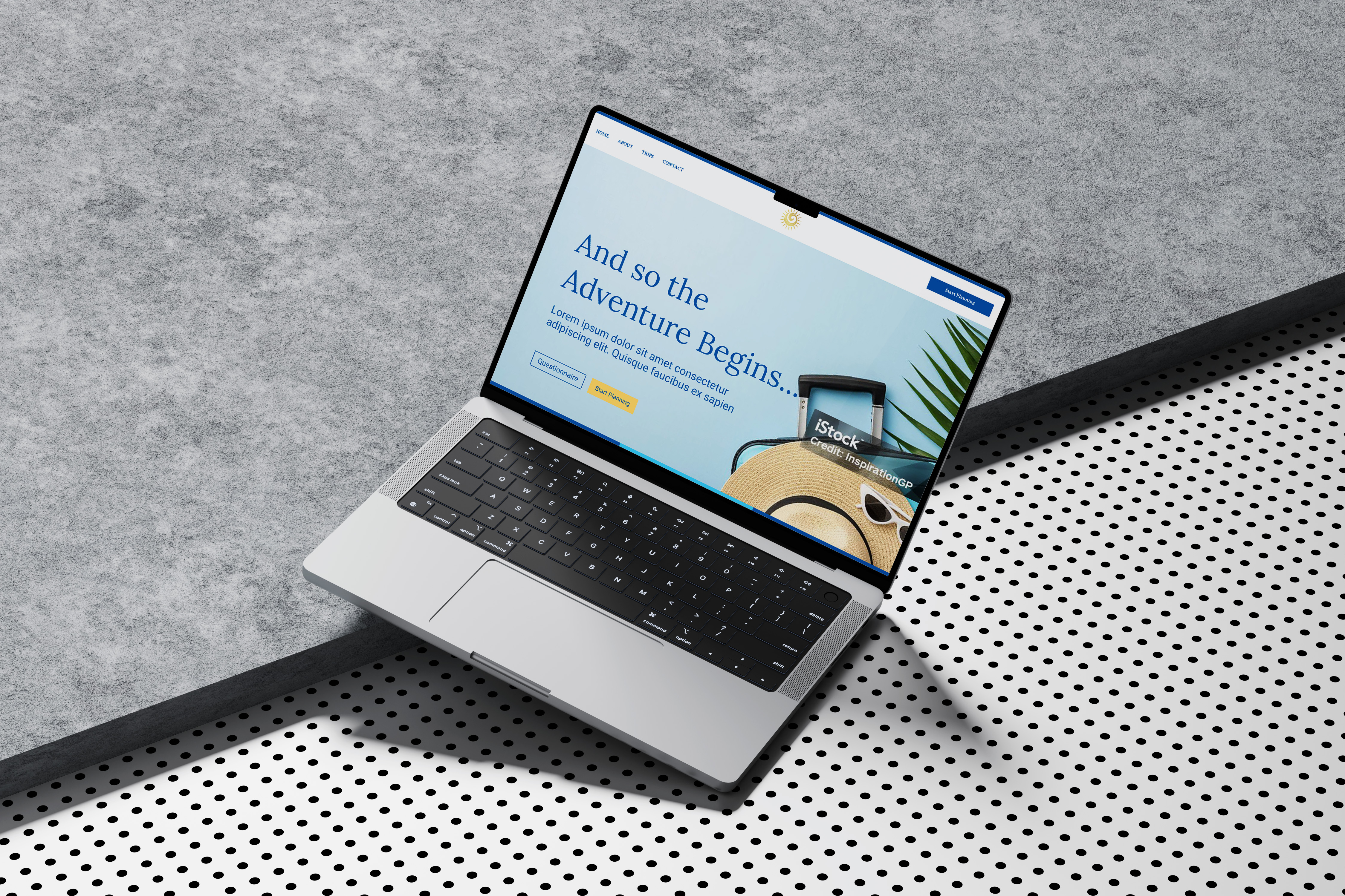 Open laptop on a textured gray surface displaying a webpage with the heading 'And so the Adventure Begins' and a travel theme including a hat, sunglasses, and a suitcase.