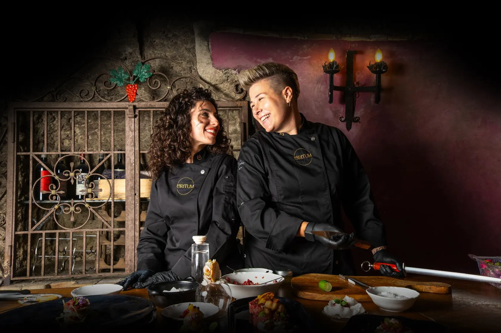 Due chef sorridenti in giacca nera da cucina con logo Oritum ridono davanti a un tavolo con piatti e ingredienti.