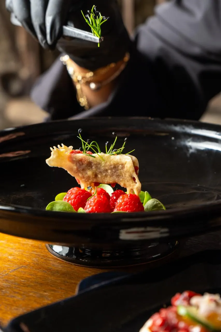 Piattino nero con dessert decorato con lamponi, foglie verdi e croccante a forma di cucciolo, mentre una mano guantata nera aggiunge un rametto decorativo.