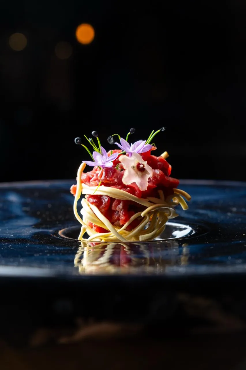 Porzione di pasta con sugo rosso guarnita con fiori commestibili su un piatto nero lucido.