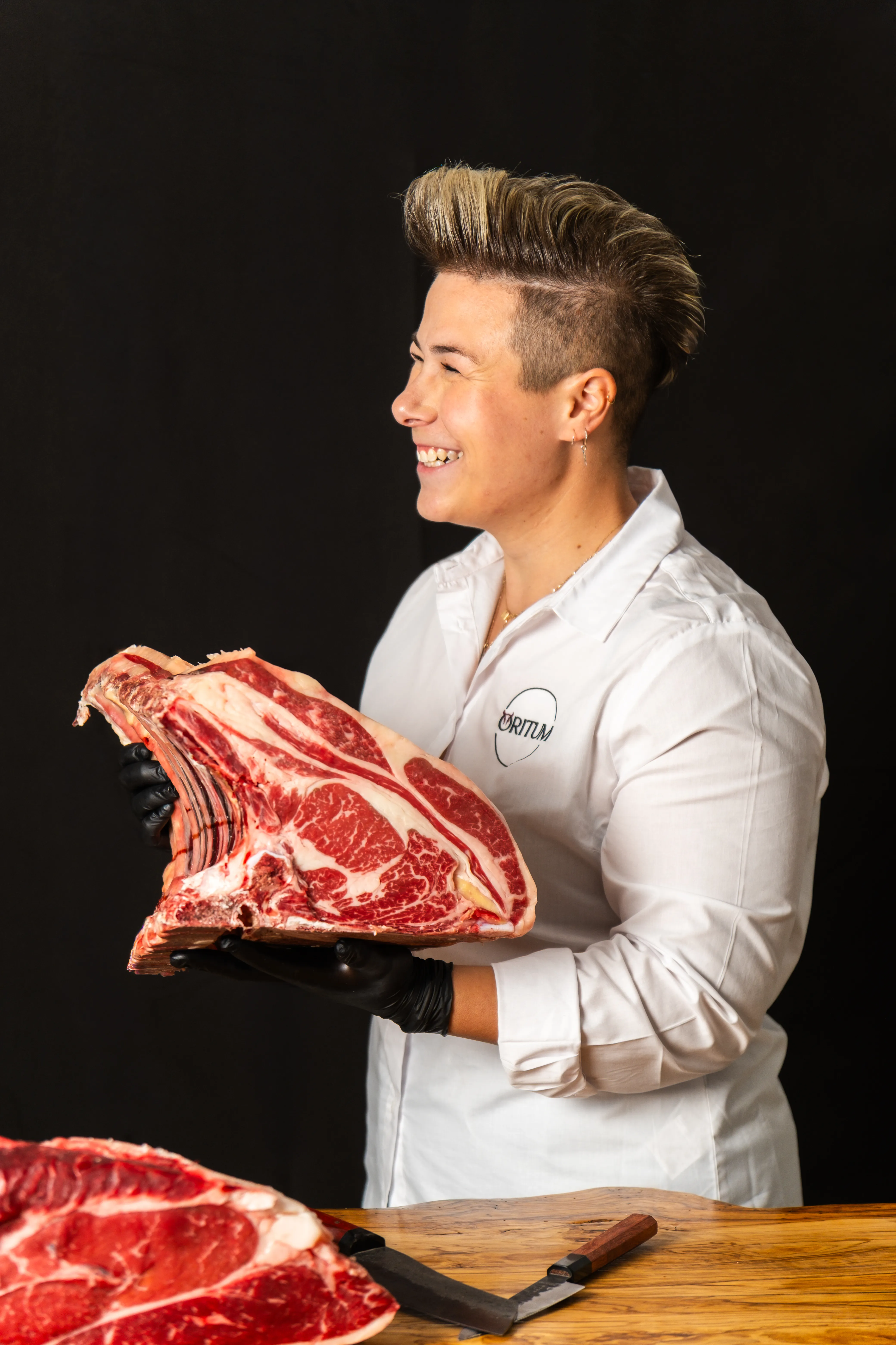 Macellaia con guanti neri che sorride mentre tiene un grande pezzo di carne su uno sfondo nero.