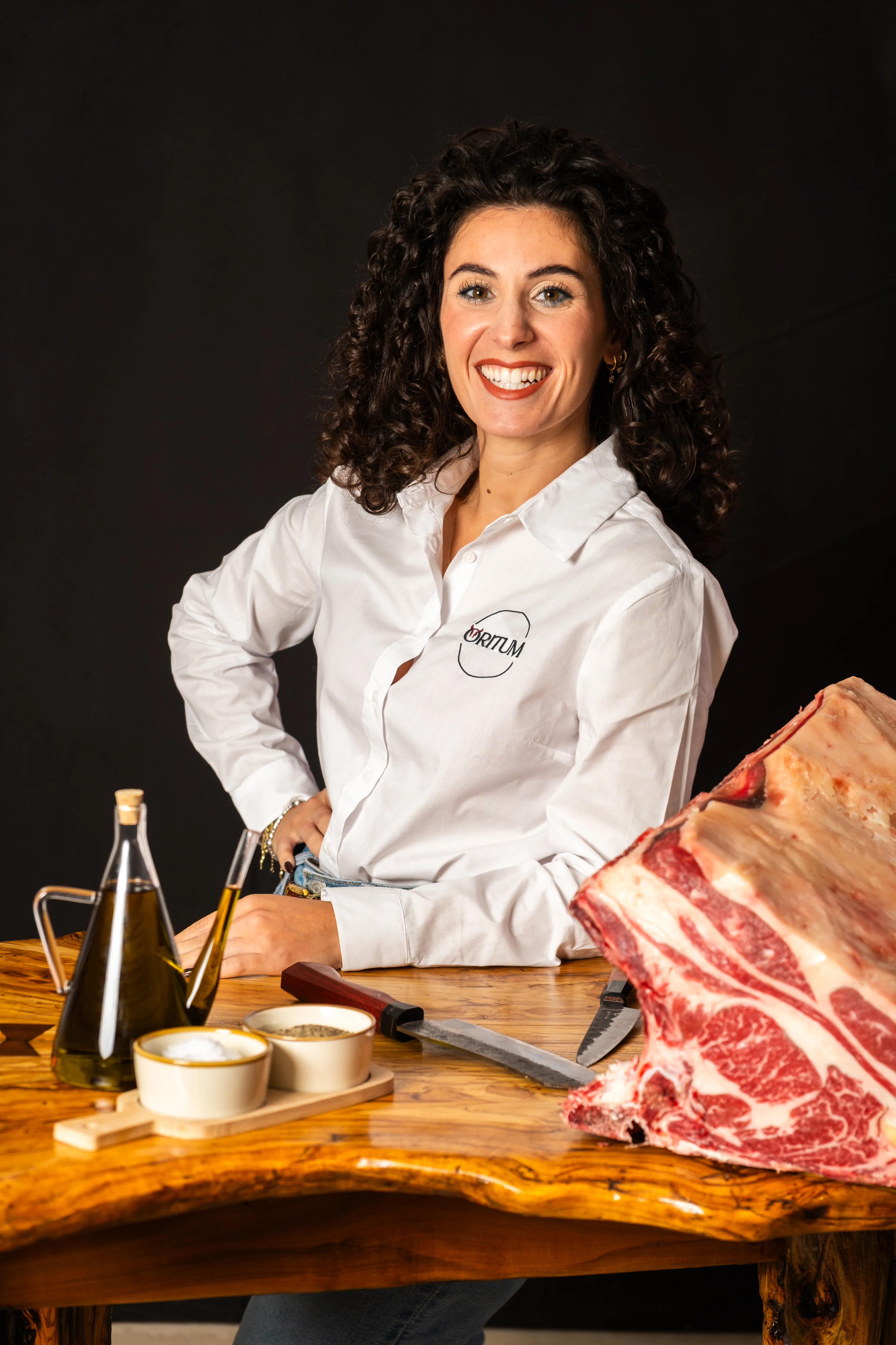 Donna sorridente con capelli ricci scuri e camicia bianca seduta a un tavolo di legno con un grande pezzo di carne cruda, olio e spezie.