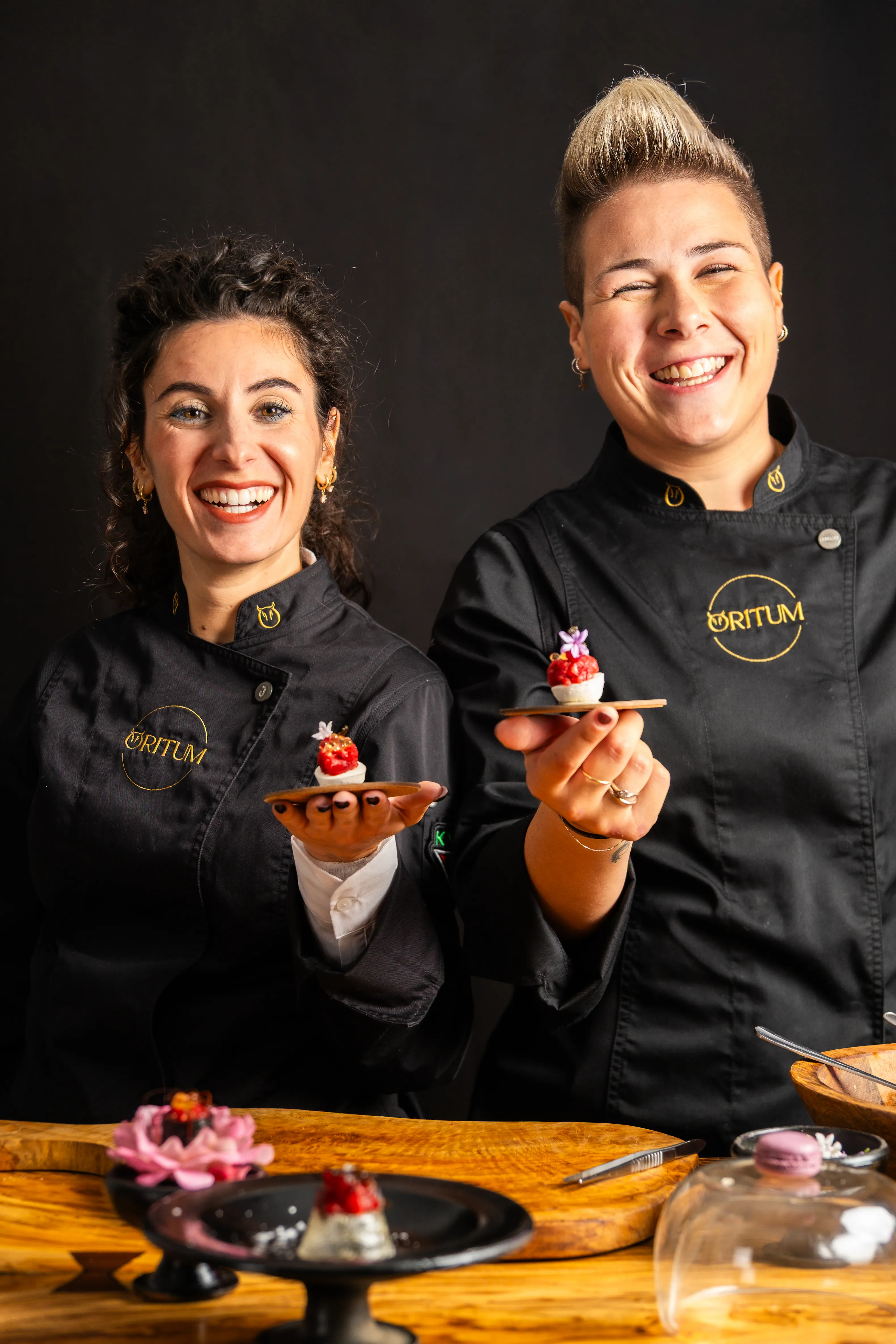 Due chef sorridenti in giacche nere con logo Oritum che mostrano dolcetti su piattini di legno davanti a sfondo nero.