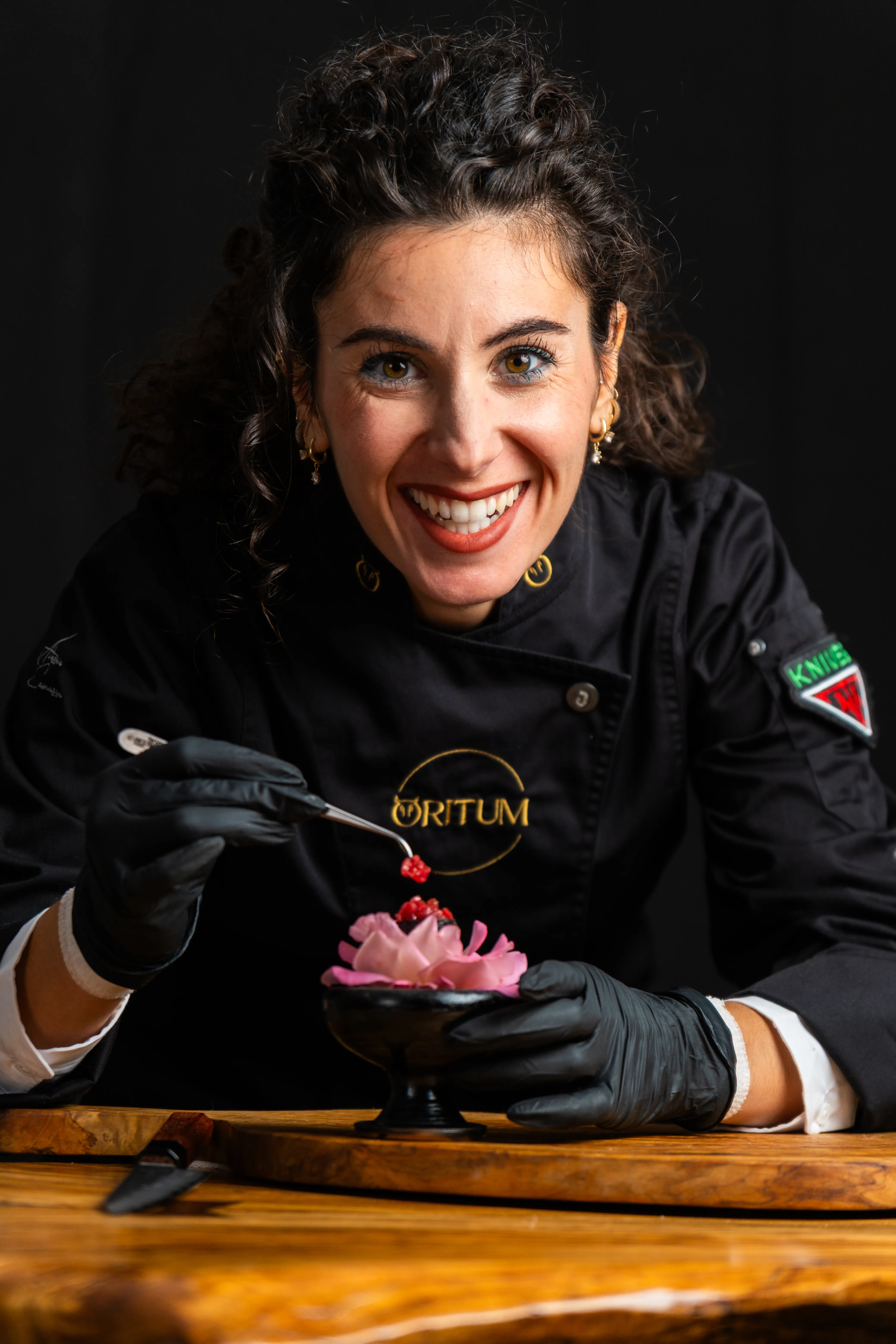 Chef sorridente con guanti neri mentre decora un piatto con fiori rosa e piccoli frutti rossi.
