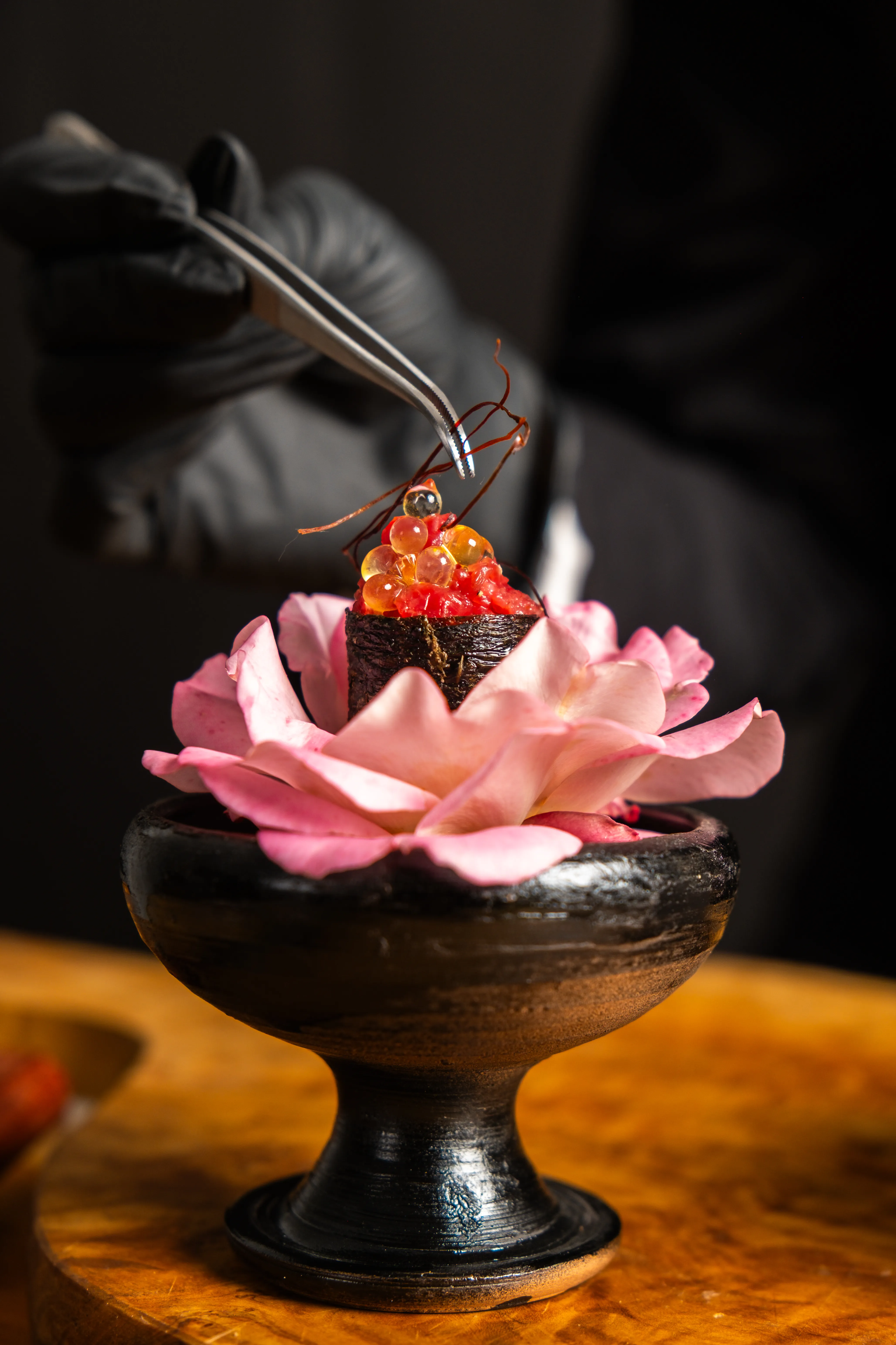 Decorazione culinaria con sushi guarnito da palline arancioni e fili sottili, presentato su un fiore rosa in un piatto nero su supporto.