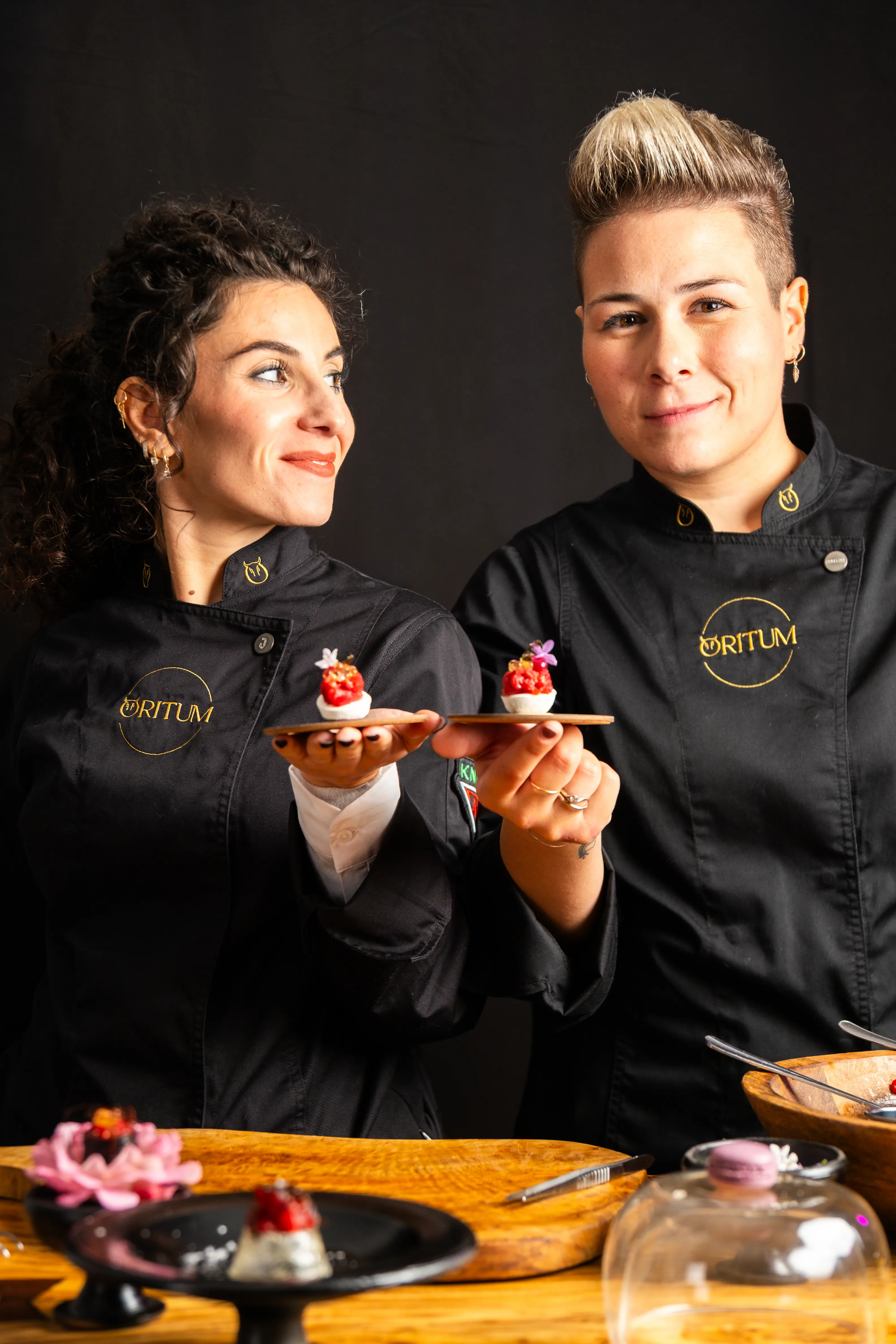 Due chef sorridenti in giacche nere con logo Oritum che mostrano dolcetti decorati su piattini di legno.