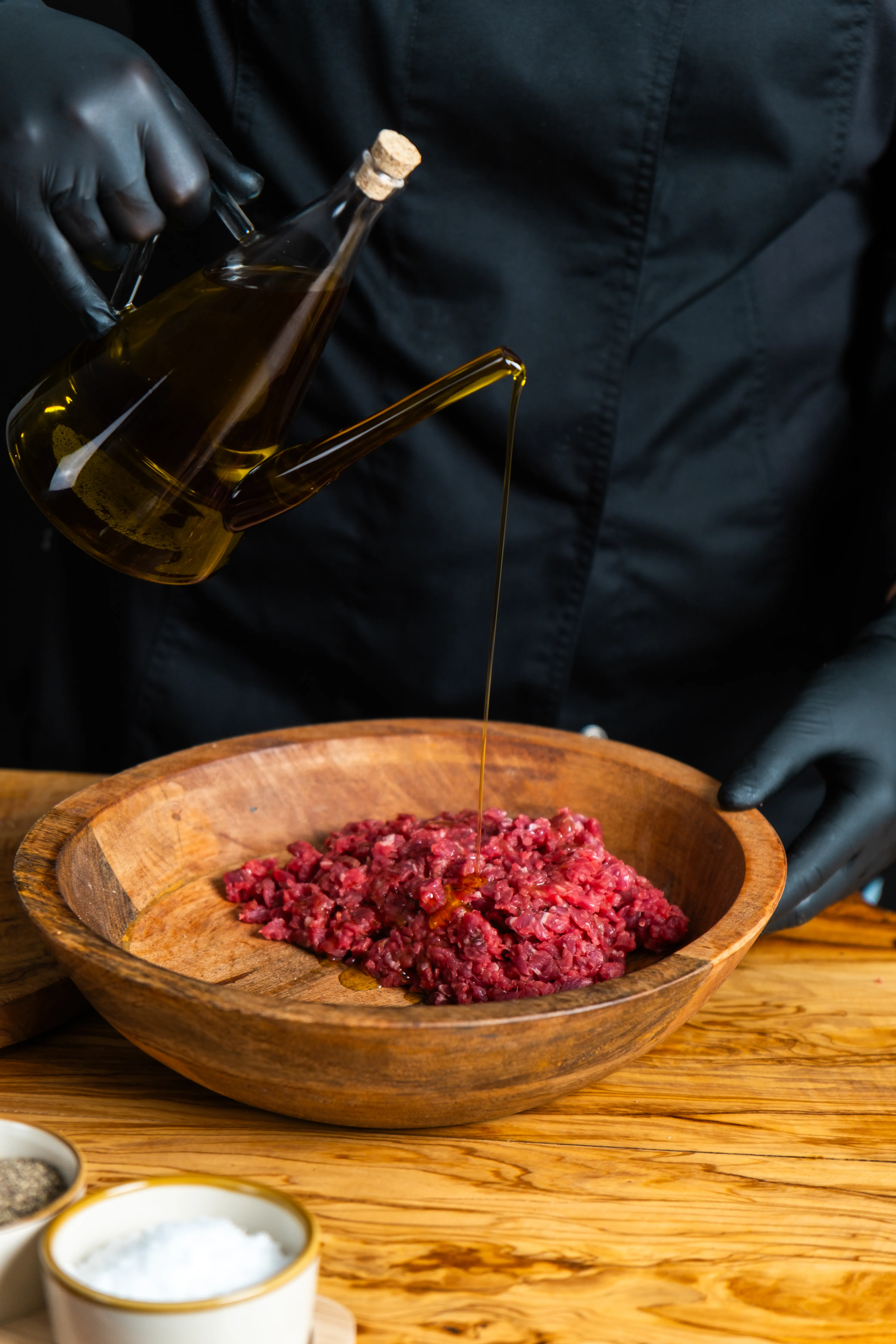Una persona con guanti neri versa olio d'oliva da una bottiglia di vetro in una ciotola di legno contenente carne macinata rossa.