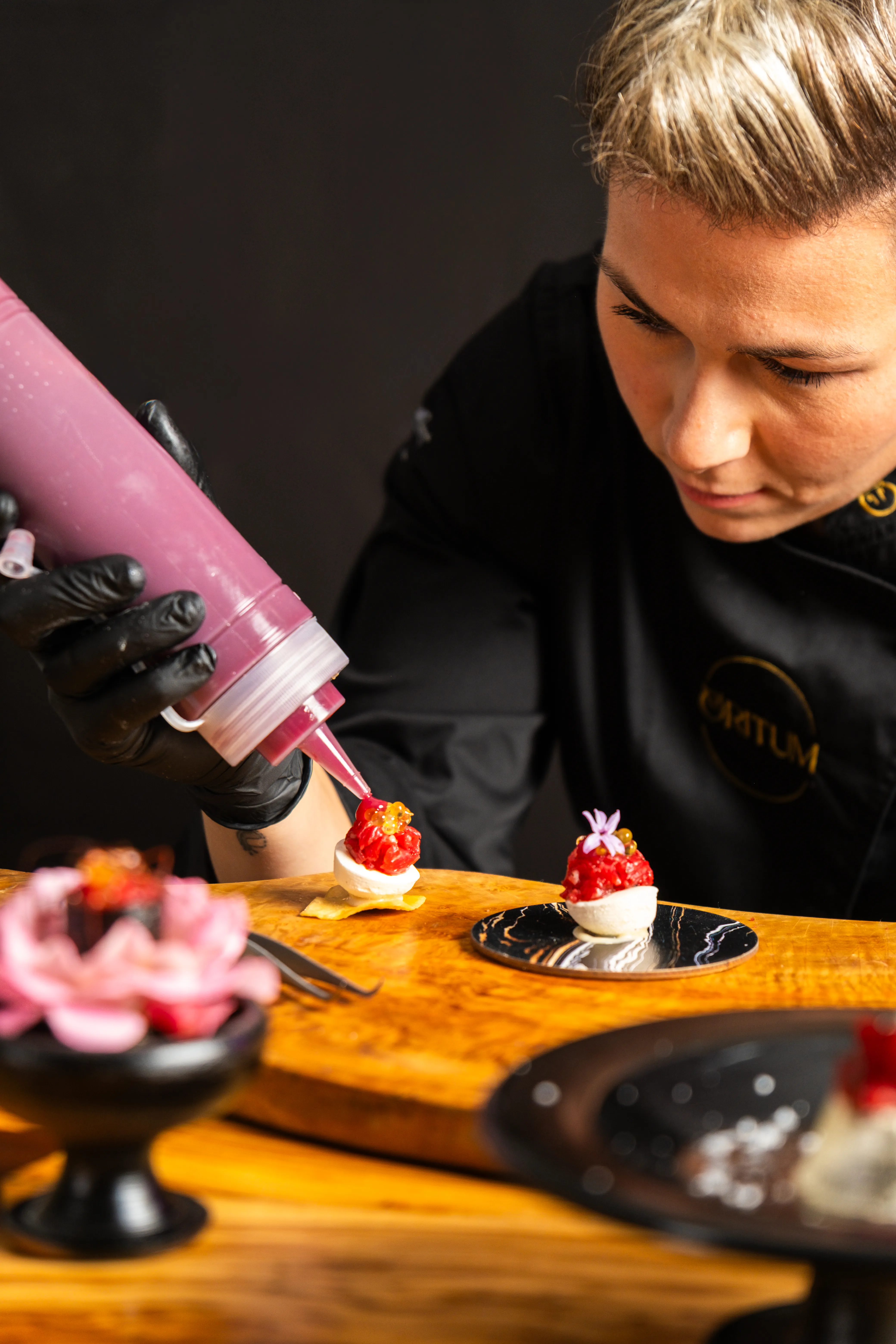 Chef con guanti neri decora un antipasto gourmet con salsa rossa su un tagliere di legno.