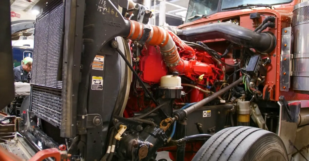 Engine repair showing heavy-duty truck with hood open, red diesel engine exposed, radiator and hoses visible in a repair shop.