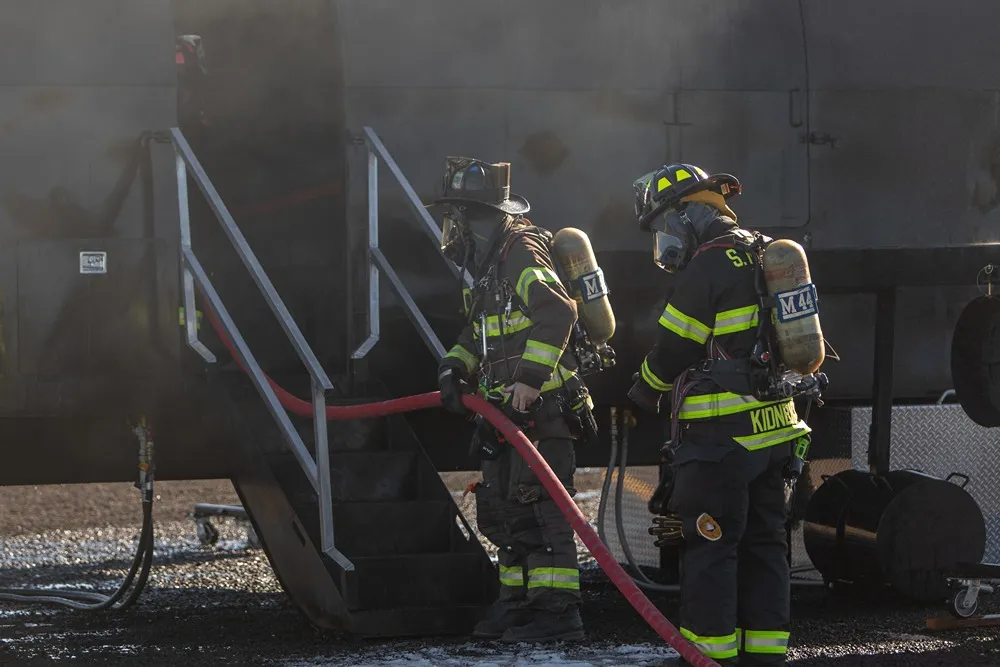 Firefighters in full gear carrying a hose into a smoking aircraft training simulator