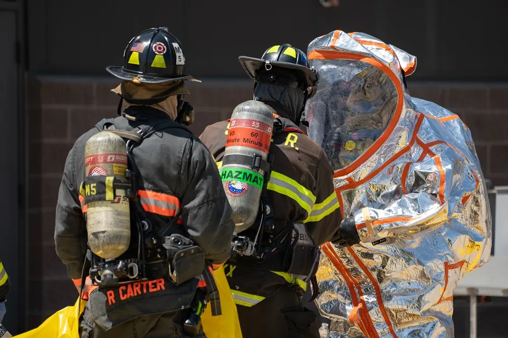 Firefighters assisting a team member in a silver hazmat suit during emergency response training