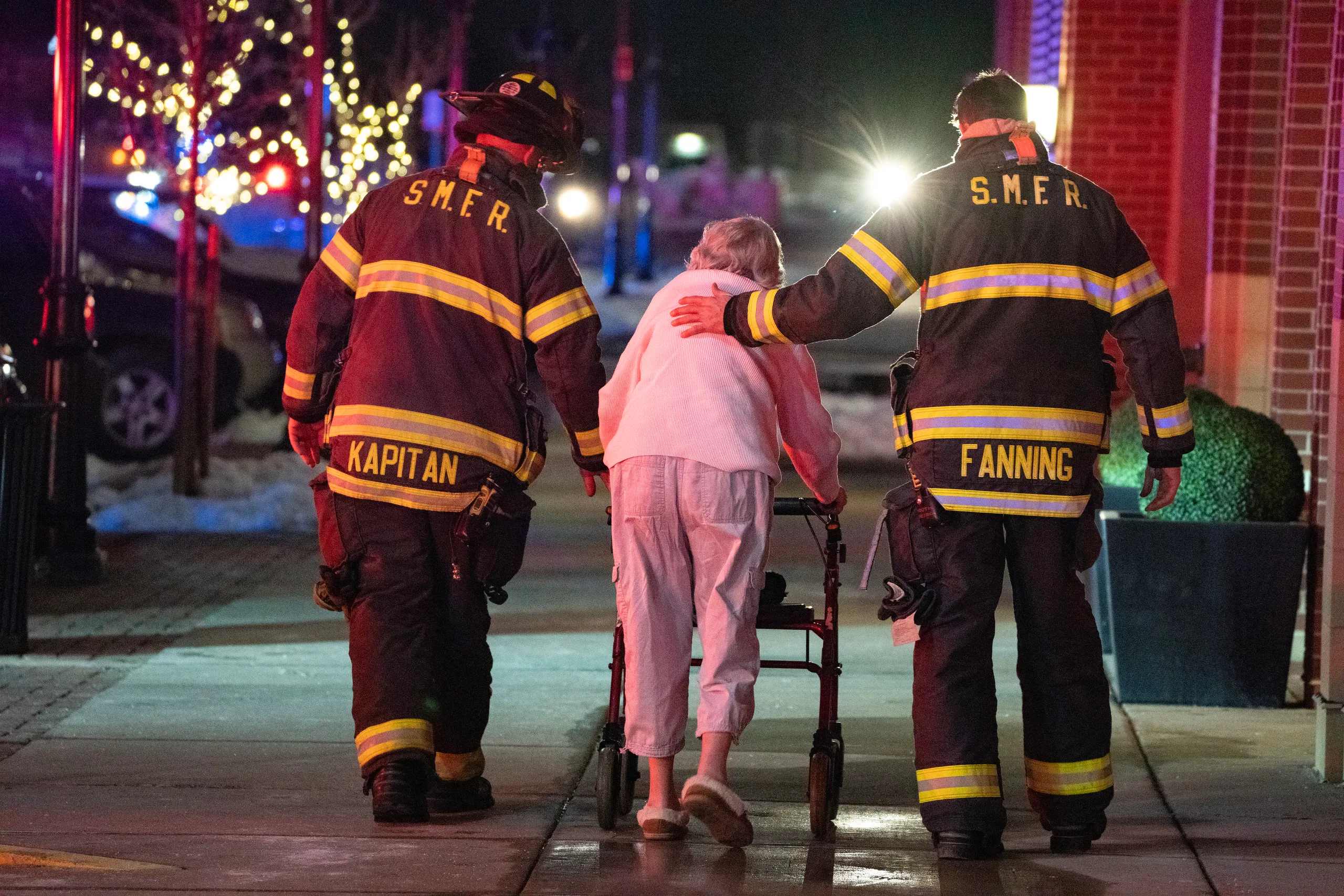 Two firefighters assisting an elderly woman with a walker during an emergency at night