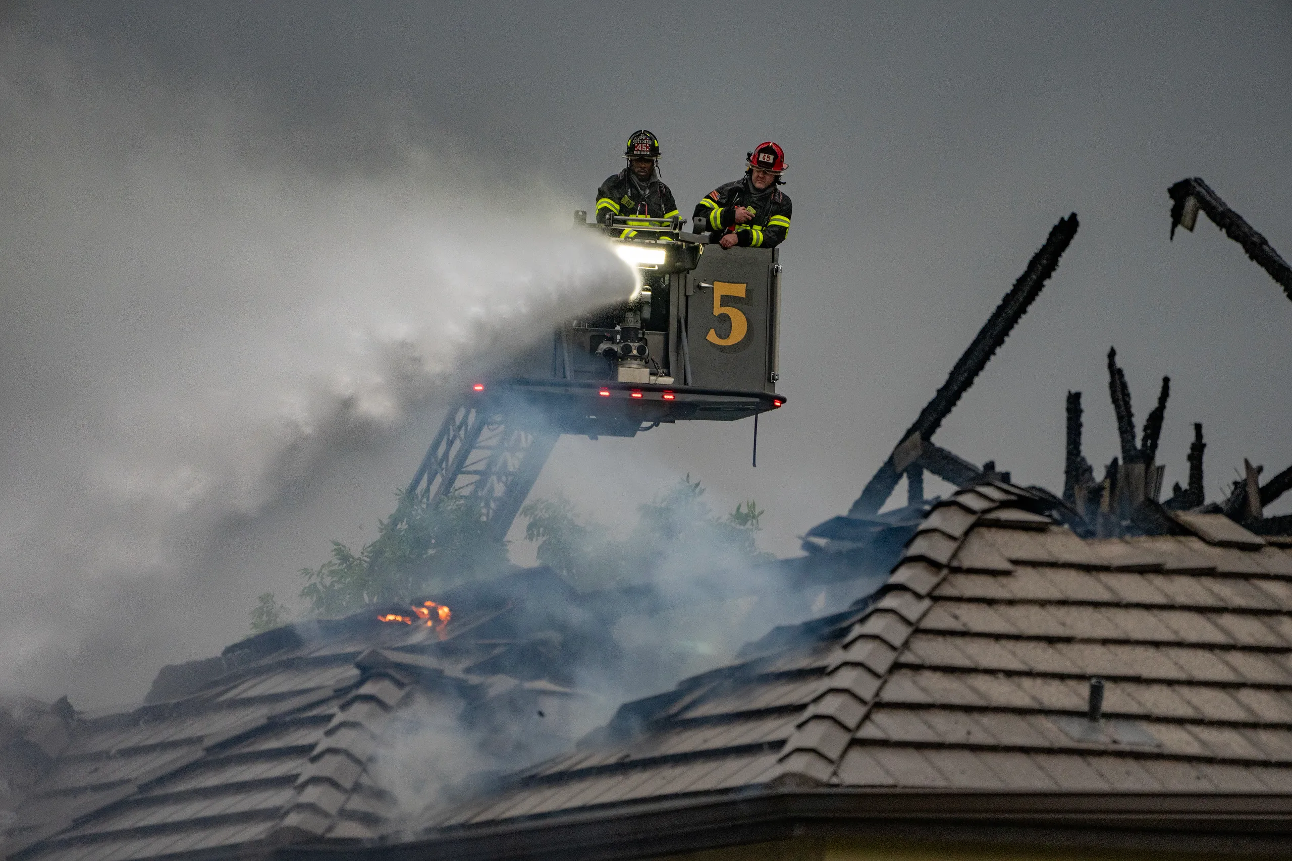 Two firefighters on a ladder truck spraying water onto a burning rooftop during a house fire
