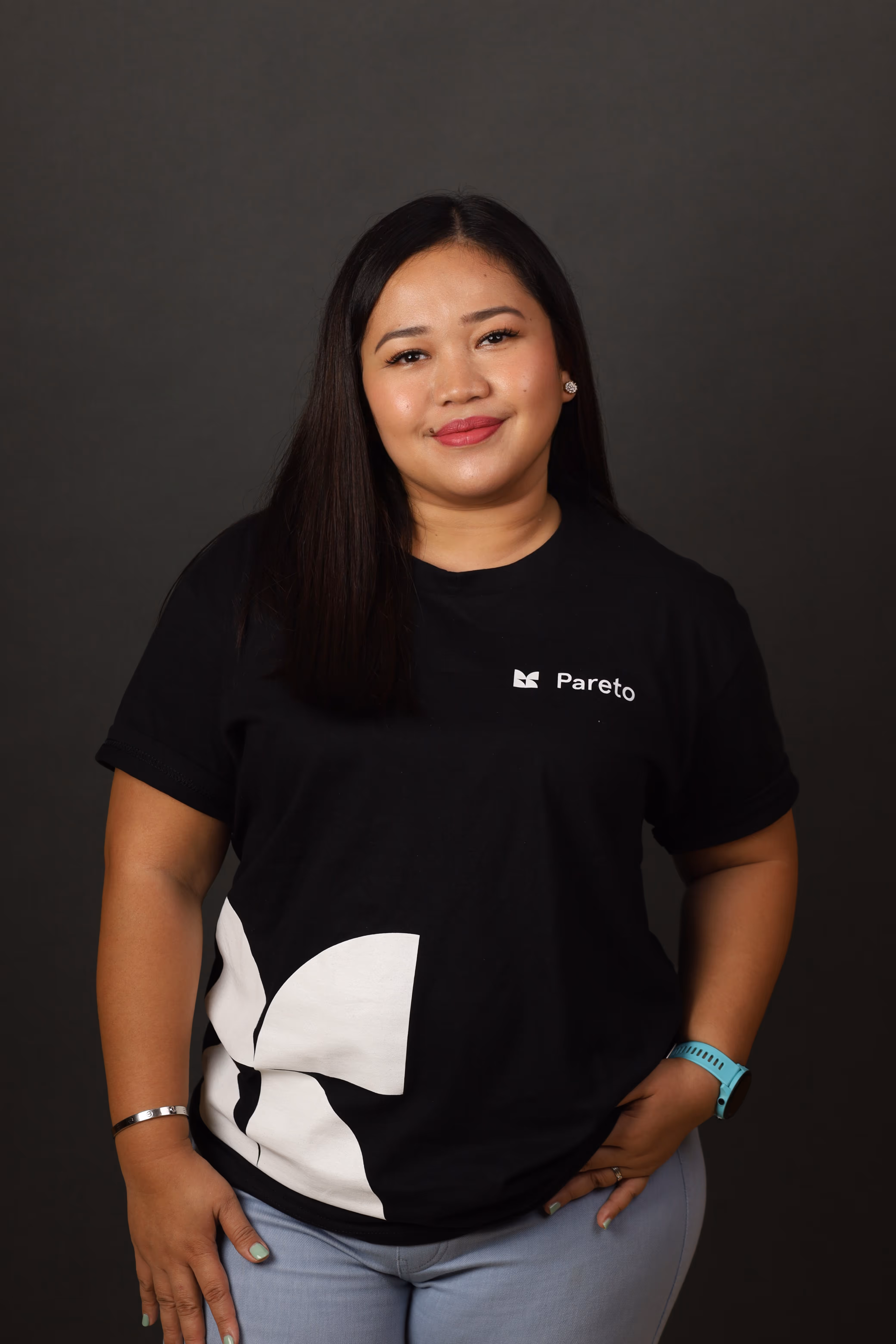 a woman poses in front of a grey backdrop with a black shirt with Pareto's logo and jeans
