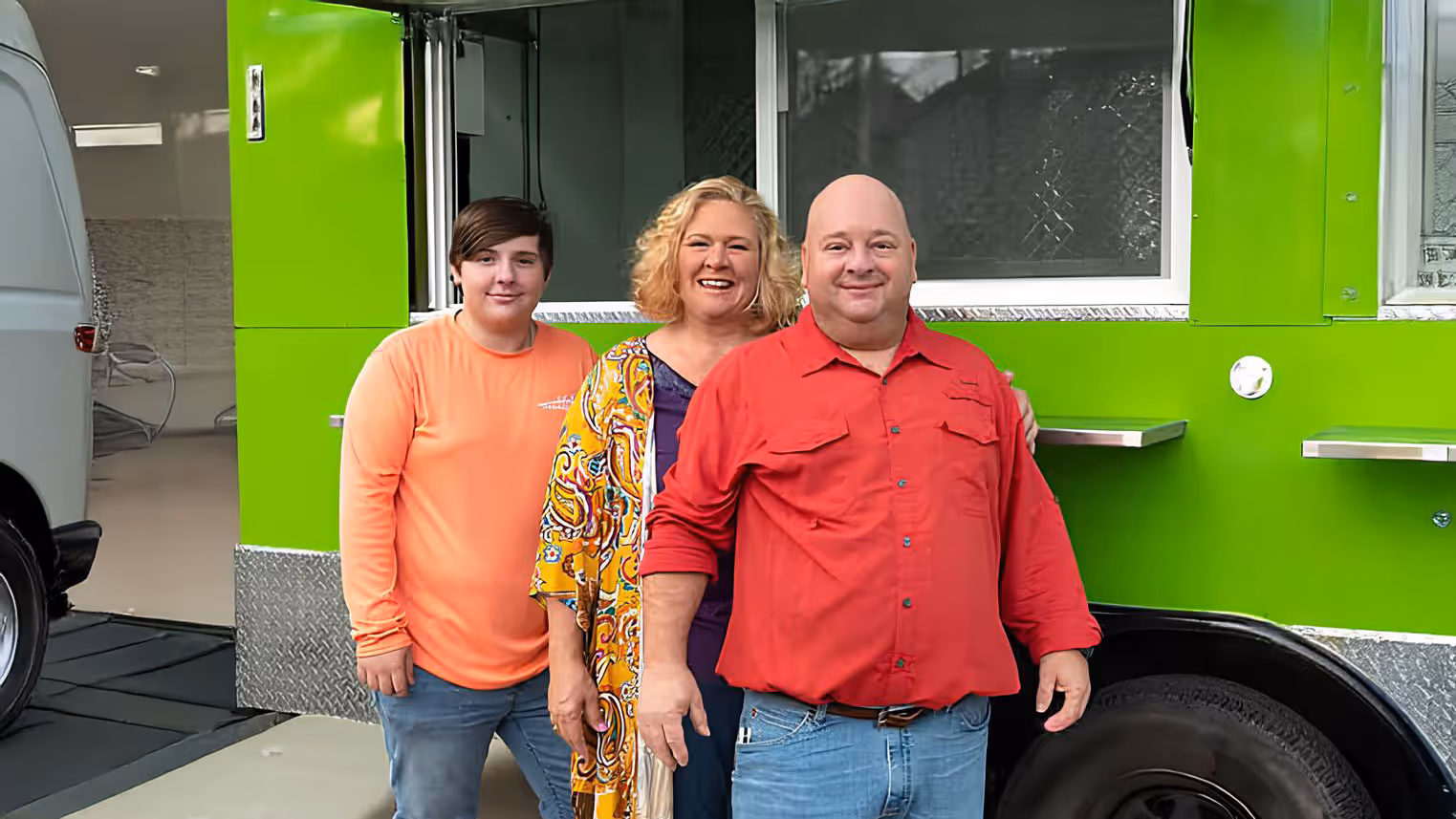 A family of three stands in front of a bright green food truck. They are smiling warmly, with casual clothing, conveying a welcoming and cheerful mood.