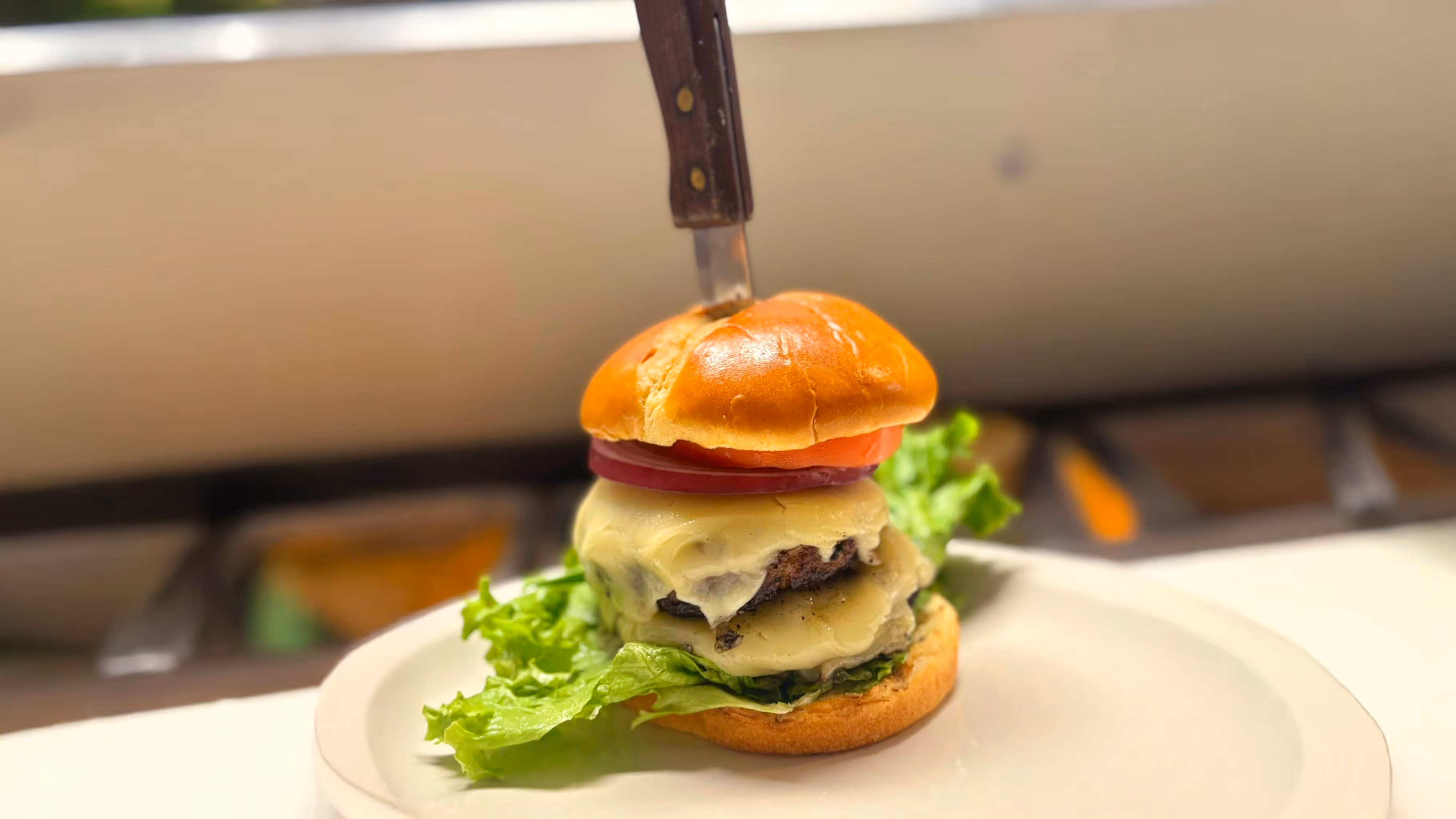 A juicy cheeseburger with lettuce, tomato, and melty cheese is skewered by a knife on a bun, set on a white plate, evoking a bold, appetizing vibe.