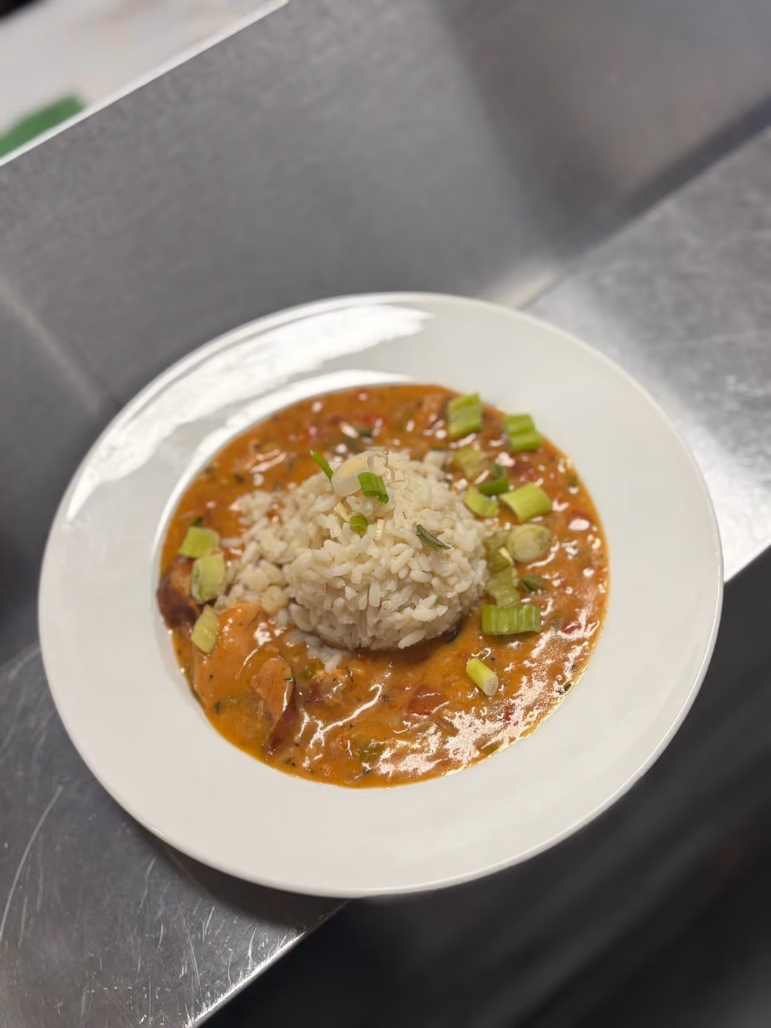 A white plate with creamy gumbo topped with chopped green onions, surrounding a mound of fluffy white rice, on a metallic surface.