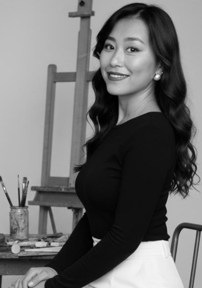 A professional studio photograph of Leah Ma happily presenting about herself. She is wearing a black top with an easel in the background. 