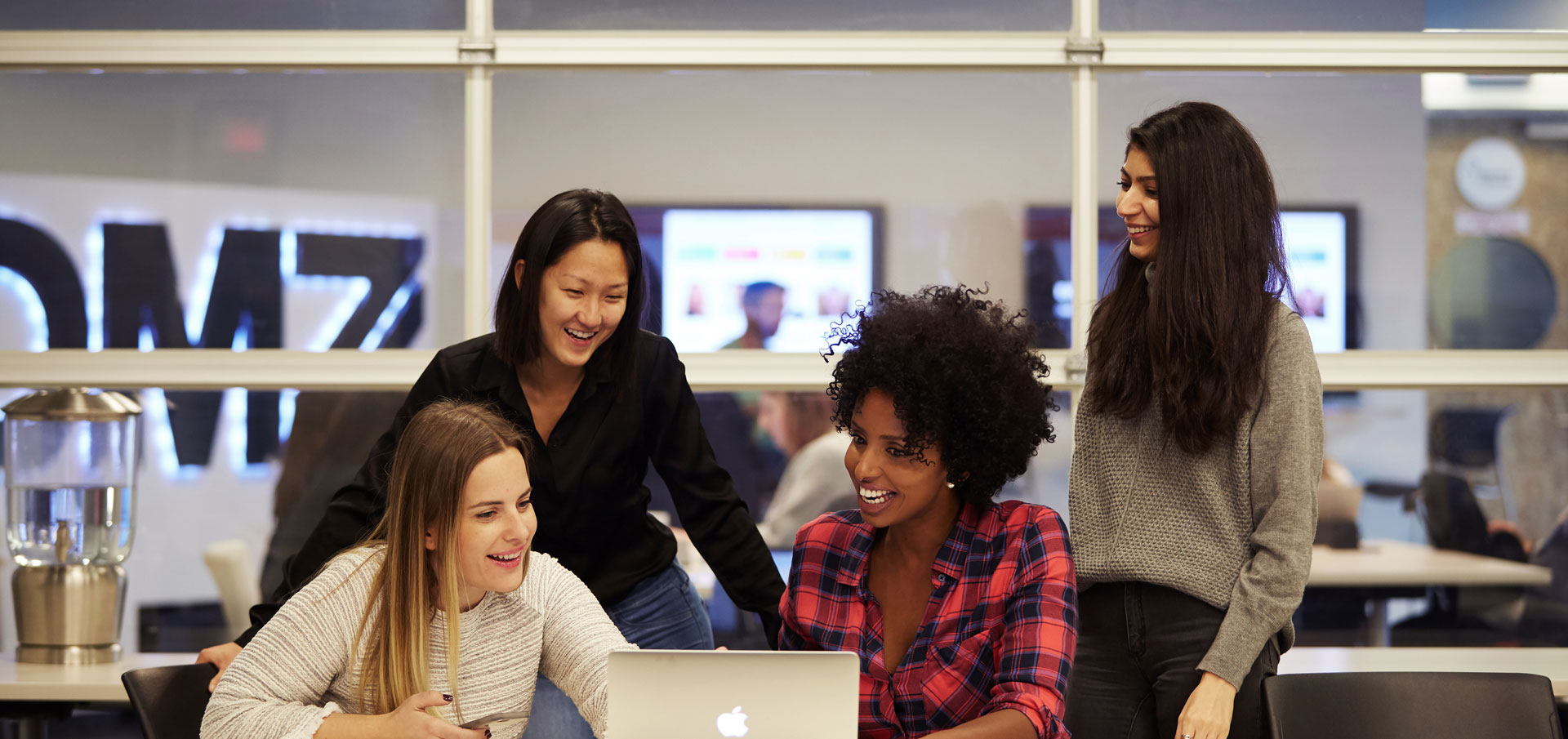 Four diverse women smiling and collaborating around a laptop in a modern office space.